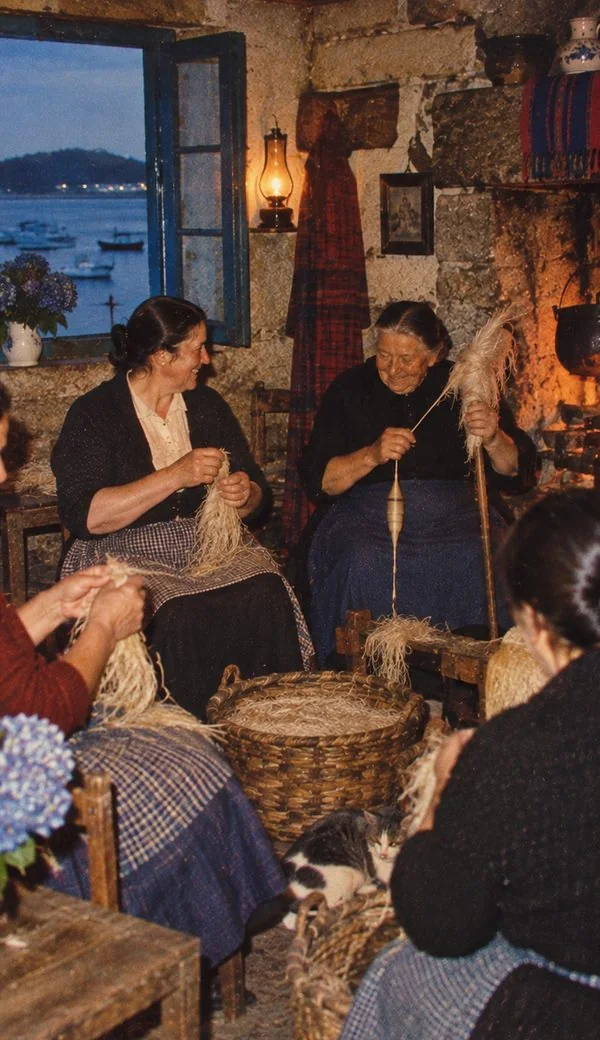 Dones filant lli vora el foc, amb la ria a l’altra banda de la finestra